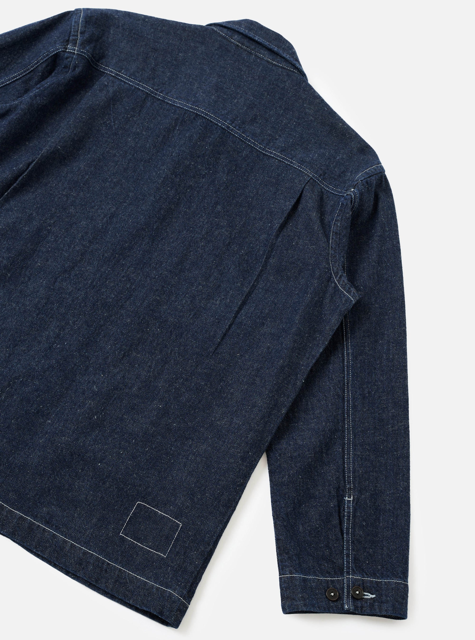 Rear three-quarter flat lay of an indigo denim troopers jacket on a white studio backdrop. Contrast white topstitching runs along the yoke, side seams, and hem. The cuff features two dark buttons. A stitched rectangular patch detail sits at the lower back hem.