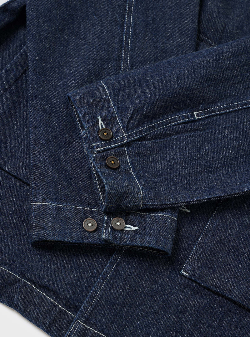 Close-up flat lay of the indigo denim Troopers Jacket on a white surface, focusing on the sleeve and cuffs. Two dark horn-style buttons fasten the placket, with two further buttons on the cuff. Contrast white topstitching runs throughout the heavy denim fabric, highlighting seam and panel construction.