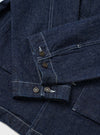 Close-up flat lay of the indigo denim Troopers Jacket on a white surface, focusing on the sleeve and cuffs. Two dark horn-style buttons fasten the placket, with two further buttons on the cuff. Contrast white topstitching runs throughout the heavy denim fabric, highlighting seam and panel construction.