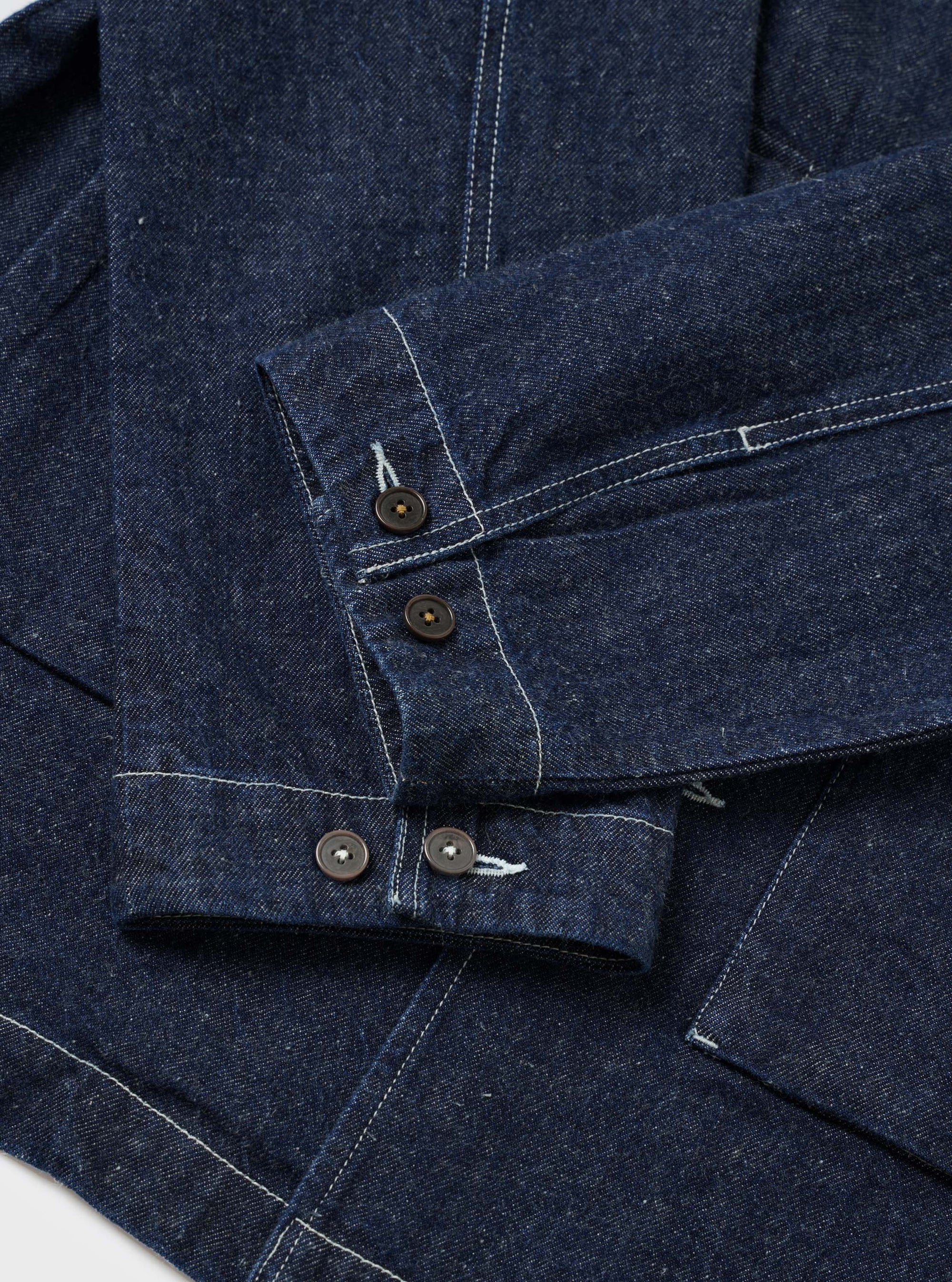Close-up flat lay of the indigo denim Troopers Jacket on a white surface, focusing on the sleeve and cuffs. Two dark horn-style buttons fasten the placket, with two further buttons on the cuff. Contrast white topstitching runs throughout the heavy denim fabric, highlighting seam and panel construction.