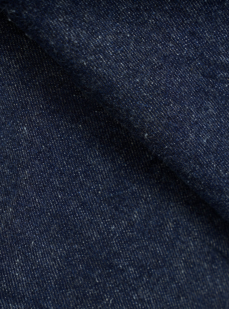 Close-up of indigo denim fabric showing a textured twill weave with flecks of white yarn throughout. The deep navy blue cloth is folded, creating soft ridges that highlight the fabric's weight and slub texture. No garment construction details are visible.