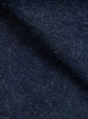 Close-up of indigo denim fabric showing a textured twill weave with flecks of white yarn throughout. The deep navy blue cloth is folded, creating soft ridges that highlight the fabric's weight and slub texture. No garment construction details are visible.
