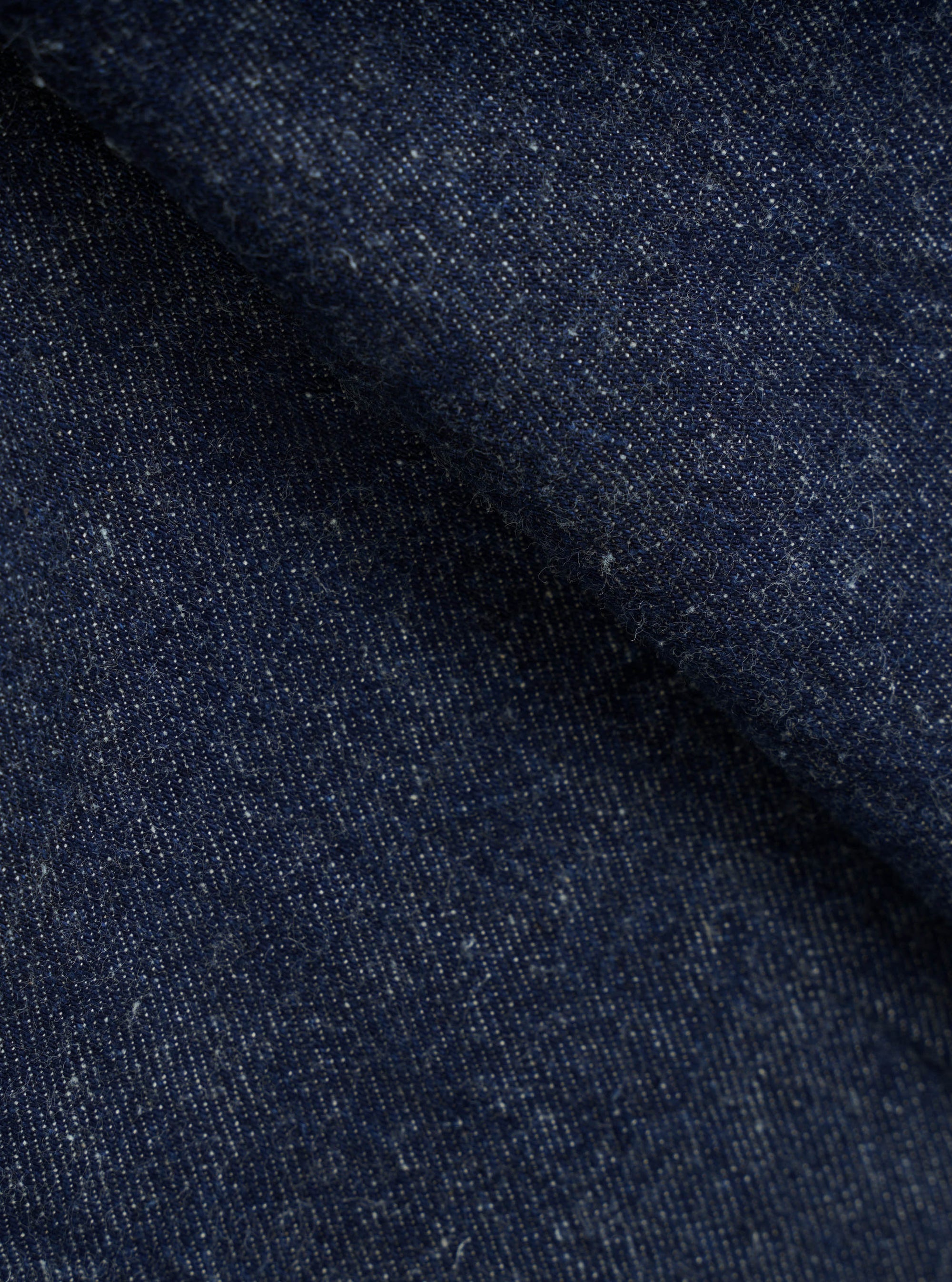 Close-up of indigo denim fabric showing a textured twill weave with flecks of white yarn throughout. The deep navy blue cloth is folded, creating soft ridges that highlight the fabric's weight and slub texture. No garment construction details are visible.