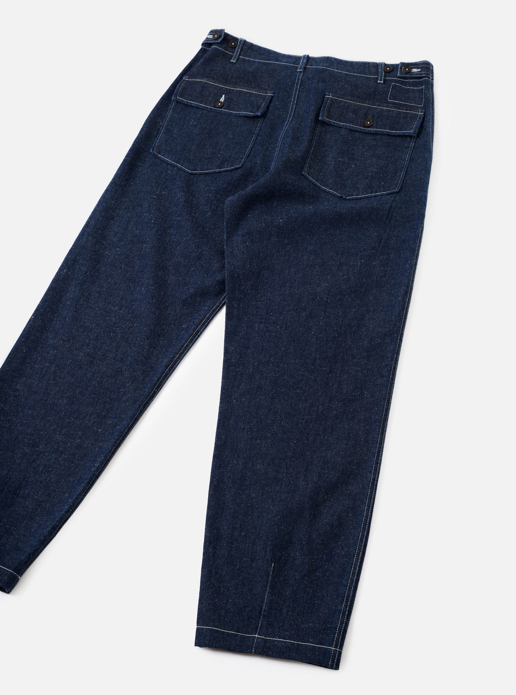 Indigo denim fatigue trousers laid flat on a light grey surface, showing the rear. Two large flap patch pockets sit at the back waist, each with a brass button closure. Contrast white topstitching runs throughout. The waistband features additional brass hardware at the sides. Wide-leg silhouette tapers slightly to a cuffed hem.