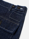 Close-up detail of indigo denim fatigue trousers on a plain light grey studio backdrop. The shot focuses on the waistband and front patch pocket area, showing two black buttons on a tab waistband closure, contrast white topstitching throughout, and a large rectangular patch pocket below.