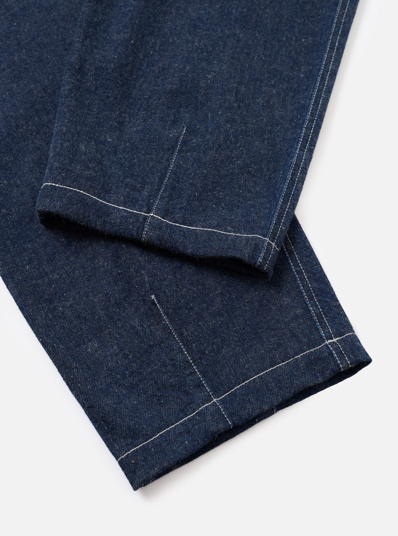 Close-up flat lay of the lower leg and hem of the Indigo Future Denim Fatigue Pant on a white surface. Dark indigo denim with a slubbed texture. Contrast white topstitching runs along the outseam and hem. A single-turn cuff is folded at the ankle, with a welt pocket opening visible above it on the leg.