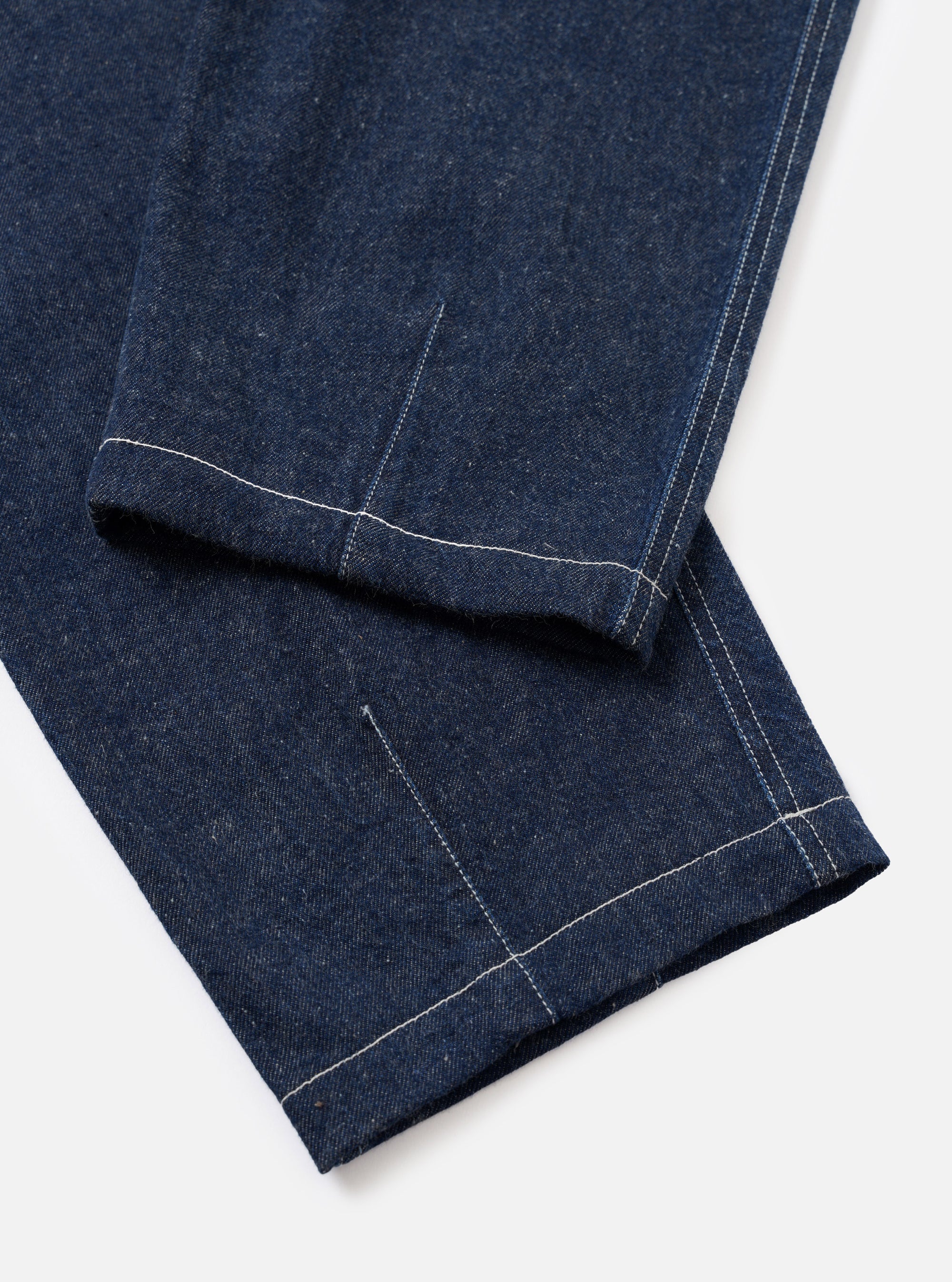 Close-up flat lay of the lower leg and hem of the Indigo Future Denim Fatigue Pant on a white surface. Dark indigo denim with a slubbed texture. Contrast white topstitching runs along the outseam and hem. A single-turn cuff is folded at the ankle, with a welt pocket opening visible above it on the leg.
