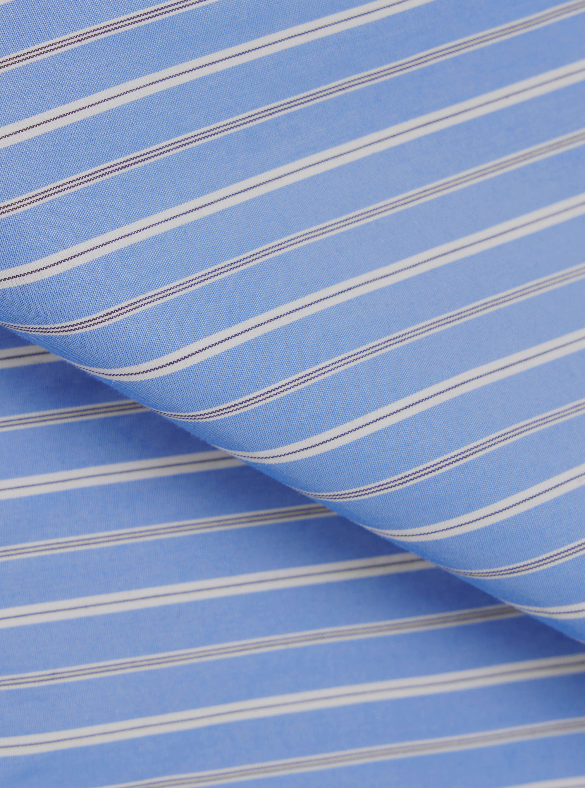 Close-up of the blue stripe compact cotton fabric used in the Lucky Shirt. The woven textile features a repeating stripe pattern of white and navy pinstripes on a mid-blue ground. The fabric is folded to show depth and drape, with a smooth, fine weave visible across the surface.