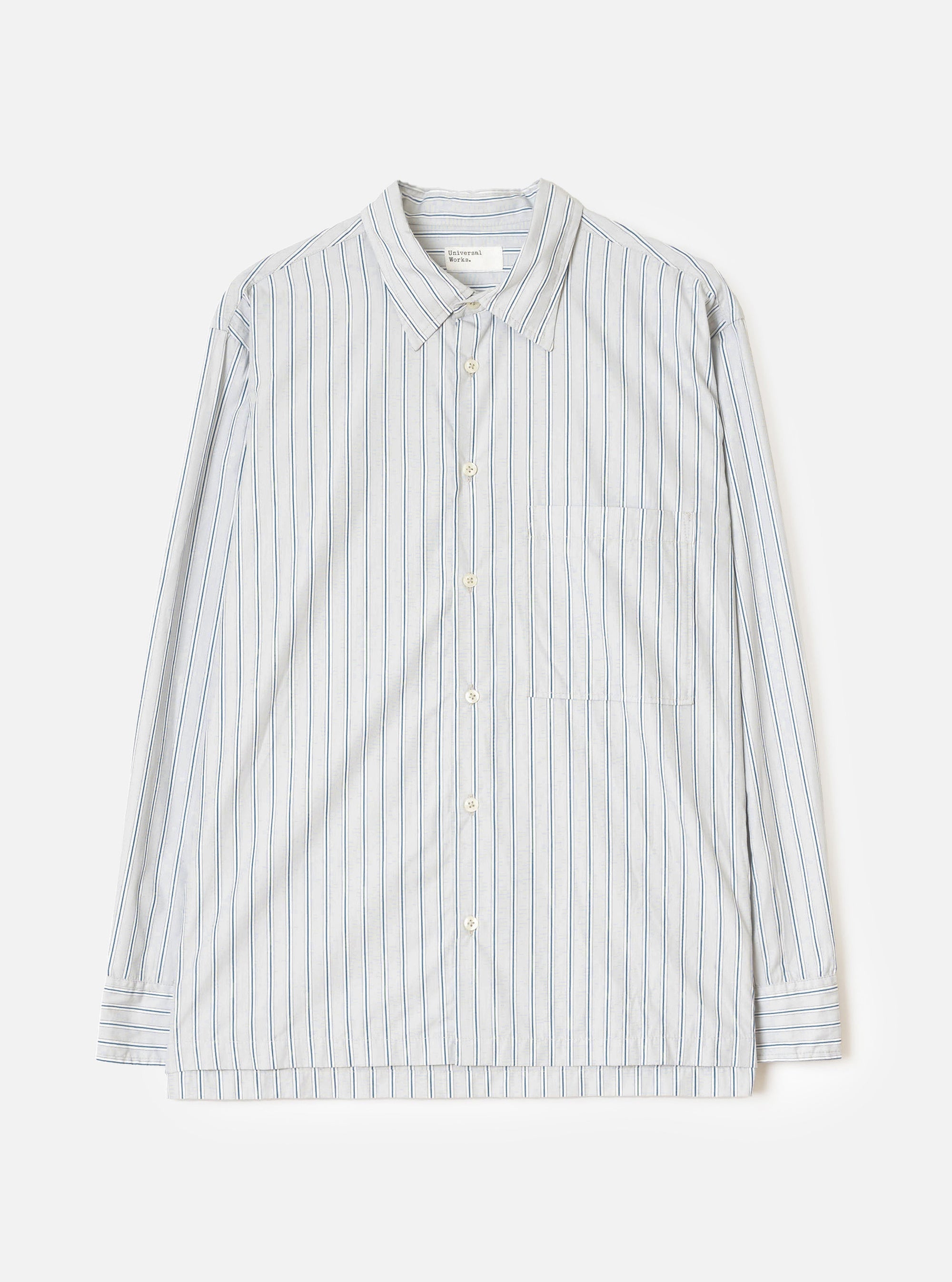 Flat lay of a grey striped long-sleeve shirt on a light grey seamless backdrop. The shirt features a rounded point collar, a concealed placket with cream buttons, a left chest patch pocket, and buttoned barrel cuffs. The fabric is a compact cotton with vertical blue and dark navy pinstripes on a grey ground. A Universal Works woven label is visible at the collar.