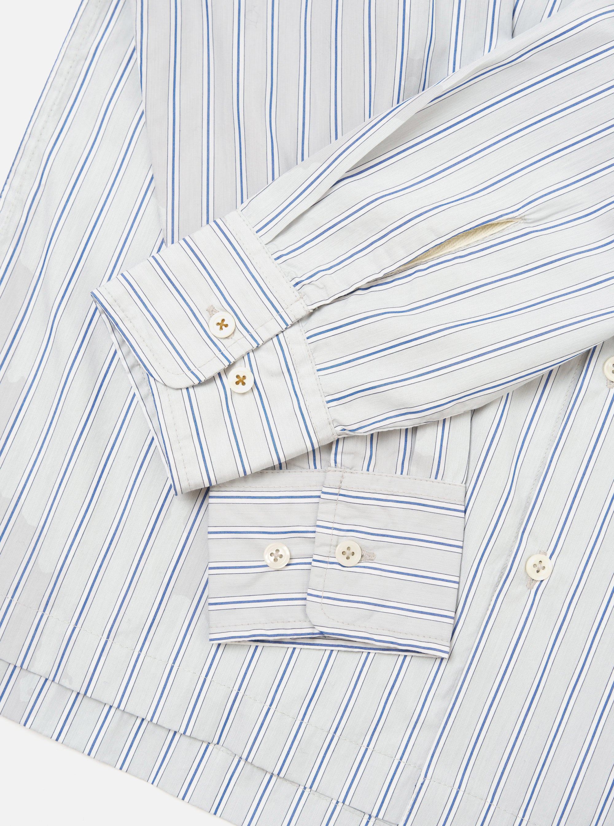 Flat lay close-up of the grey striped Lucky Shirt on a white surface, focusing on the cuffs and lower sleeves. The compact cotton fabric features blue and navy vertical stripes on a light grey ground. Both barrel cuffs are unfolded and fastened with two cream four-hole buttons each.