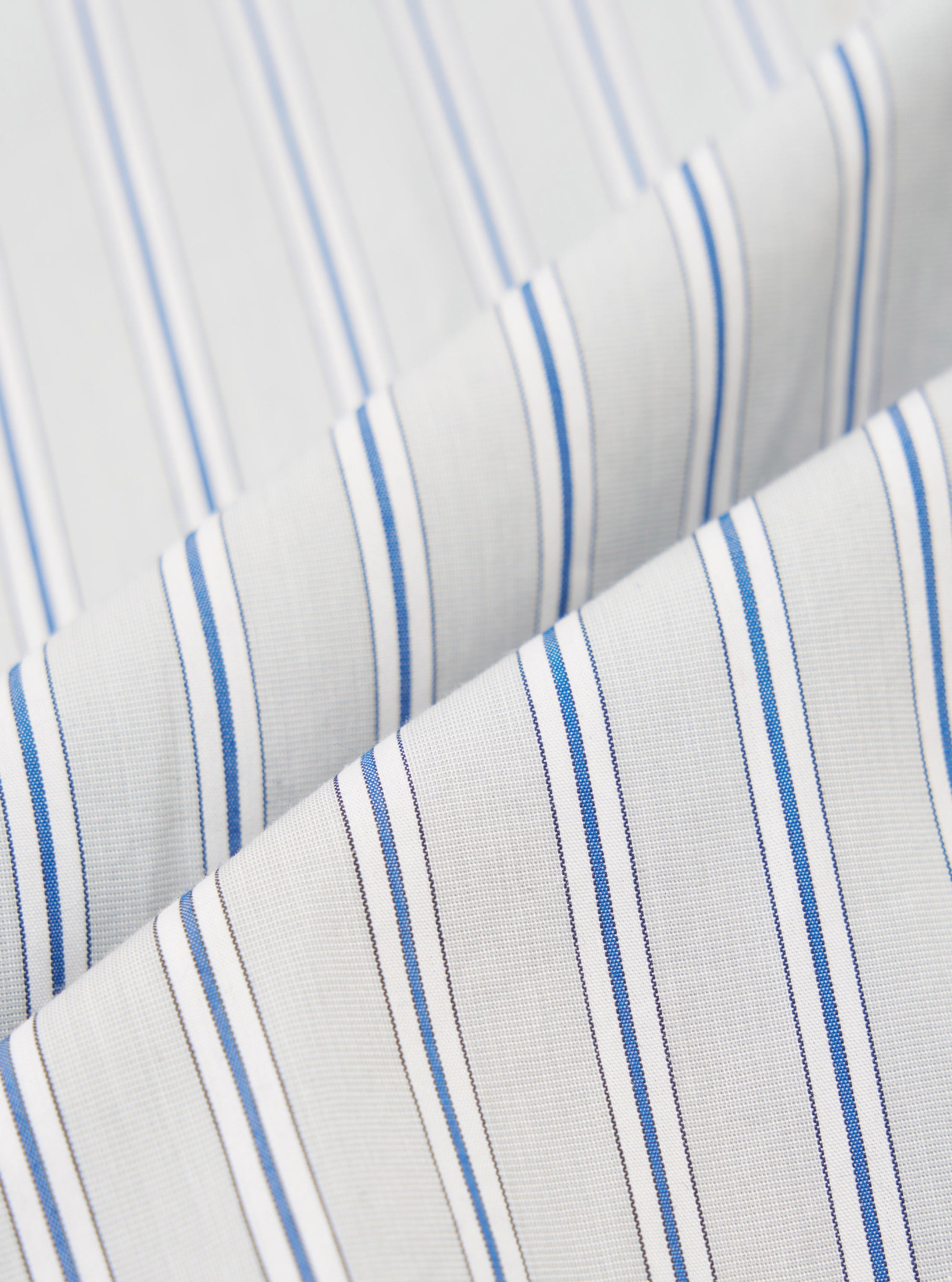 Close-up fabric shot of the grey stripe compact cotton used in the Lucky Shirt. Two folded layers of cloth overlap diagonally, showing a white base with vertical blue and grey pinstripes in a repeating grouped pattern. The fine woven texture is clearly visible against a plain white background.