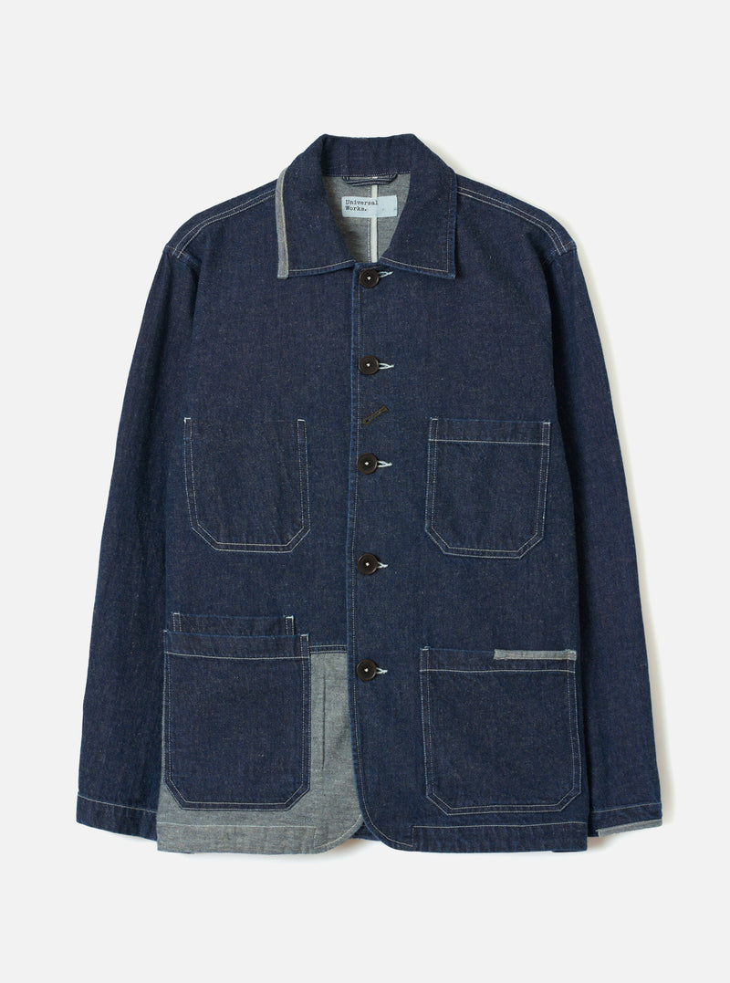 Indigo denim chore jacket on a plain light grey studio backdrop, displayed flat without a model or mannequin. Features a spread collar with grey chambray reverse, five dark buttons down the centre front, and contrast white topstitching throughout. Four large patch pockets, two chest and two lower, with the grey chambray lining visible at the hem and collar.