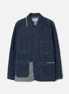 Indigo denim chore jacket on a plain light grey studio backdrop, displayed flat without a model or mannequin. Features a spread collar with grey chambray reverse, five dark buttons down the centre front, and contrast white topstitching throughout. Four large patch pockets, two chest and two lower, with the grey chambray lining visible at the hem and collar.