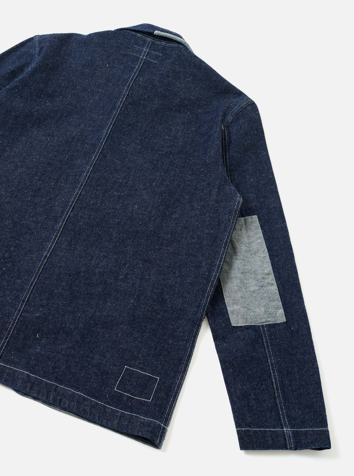 Back view of an indigo denim chore jacket, flat lay on a white surface. Contrast white topstitching runs along the back seams and hem. The right sleeve features a large rectangular elbow patch in lighter grey selvedge denim. A stitched rectangular outline sits at the lower back hem. Ribbed cuff visible at the sleeve end.