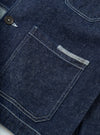 Close-up detail shot of the indigo future denim OG bakers chore jacket on a white surface. Dark indigo denim with visible twill weave texture. A large patch chest pocket features contrast white topstitching and a small selvedge tab detail. A dark horn button is visible to the left. A second patch pocket is partially visible at top right.
