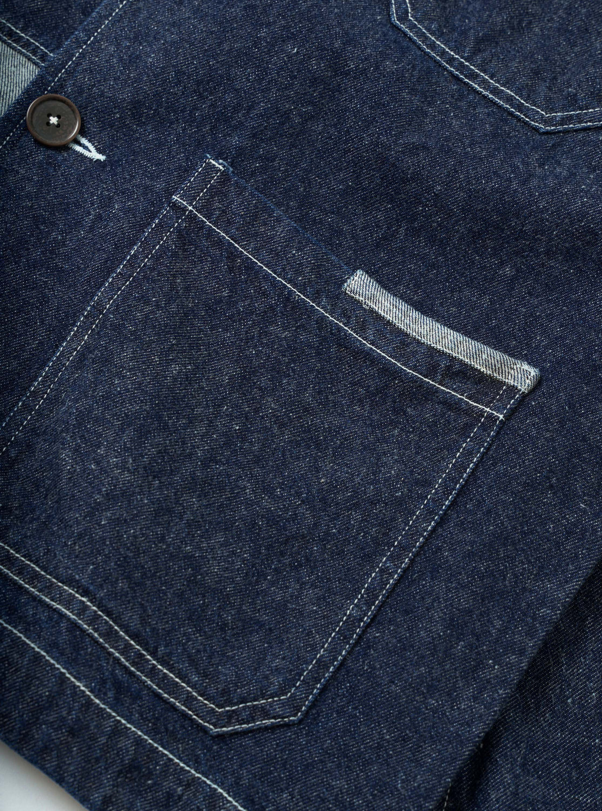 Close-up detail shot of the indigo future denim OG bakers chore jacket on a white surface. Dark indigo denim with visible twill weave texture. A large patch chest pocket features contrast white topstitching and a small selvedge tab detail. A dark horn button is visible to the left. A second patch pocket is partially visible at top right.