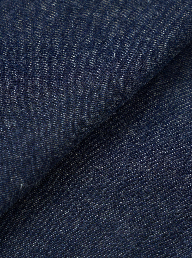 Close-up of the indigo denim fabric used in the OG Bakers Chore Jacket. The textured weave shows a deep navy-blue base with flecks of white and cream throughout, giving a slubby, neppy character. The fabric is folded, creating a diagonal crease across the frame.