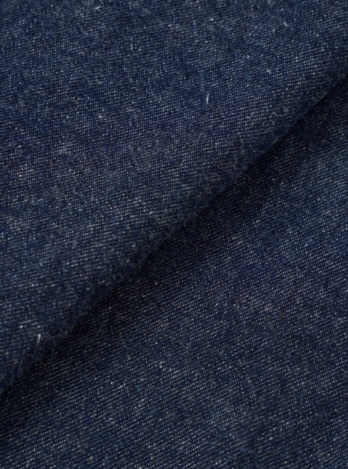 Close-up of the indigo denim fabric used in the OG Bakers Chore Jacket. The textured weave shows a deep navy-blue base with flecks of white and cream throughout, giving a slubby, neppy character. The fabric is folded, creating a diagonal crease across the frame.