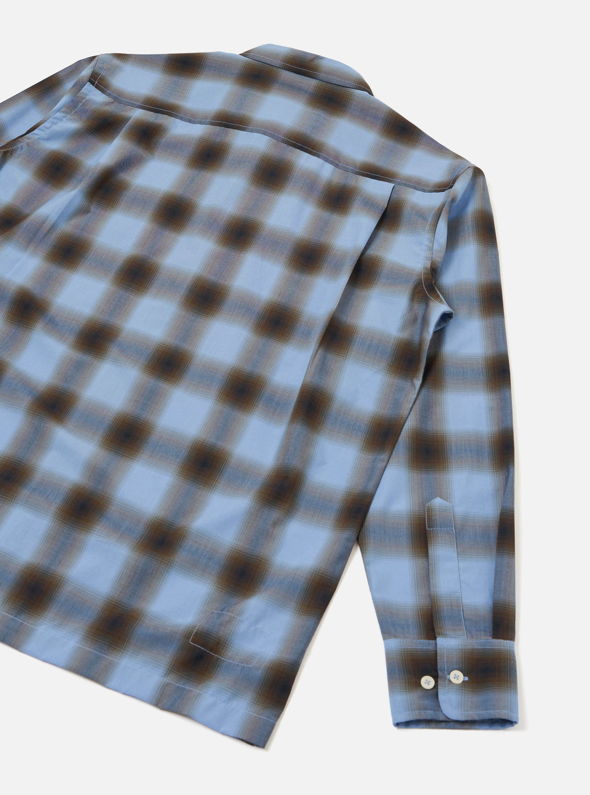 Blue check long-sleeve shirt in a soft ombre plaid with brown and dark navy tones on a light blue ground. Shot flat on a white studio backdrop, angled to show the back and one sleeve. A camp collar is visible at the top. The cuff features two white buttons and a gauntlet placket.