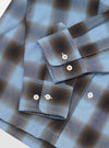 Close-up flat lay of the blue and brown check Attendant shirt on a white surface, focusing on both cuffs. Each barrel cuff fastens with two cream four-hole buttons. The woven check fabric shows a large-scale plaid in sky blue and earthy brown tones with fine white overcheck lines.