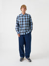 A model wearing the Universal Works Blue Check Charlie Check Attendant Shirt, viewed from front. The long-sleeve shirt features a blue check pattern, flat collar, central button closure, and two large chest flap pockets against a plain backdrop.