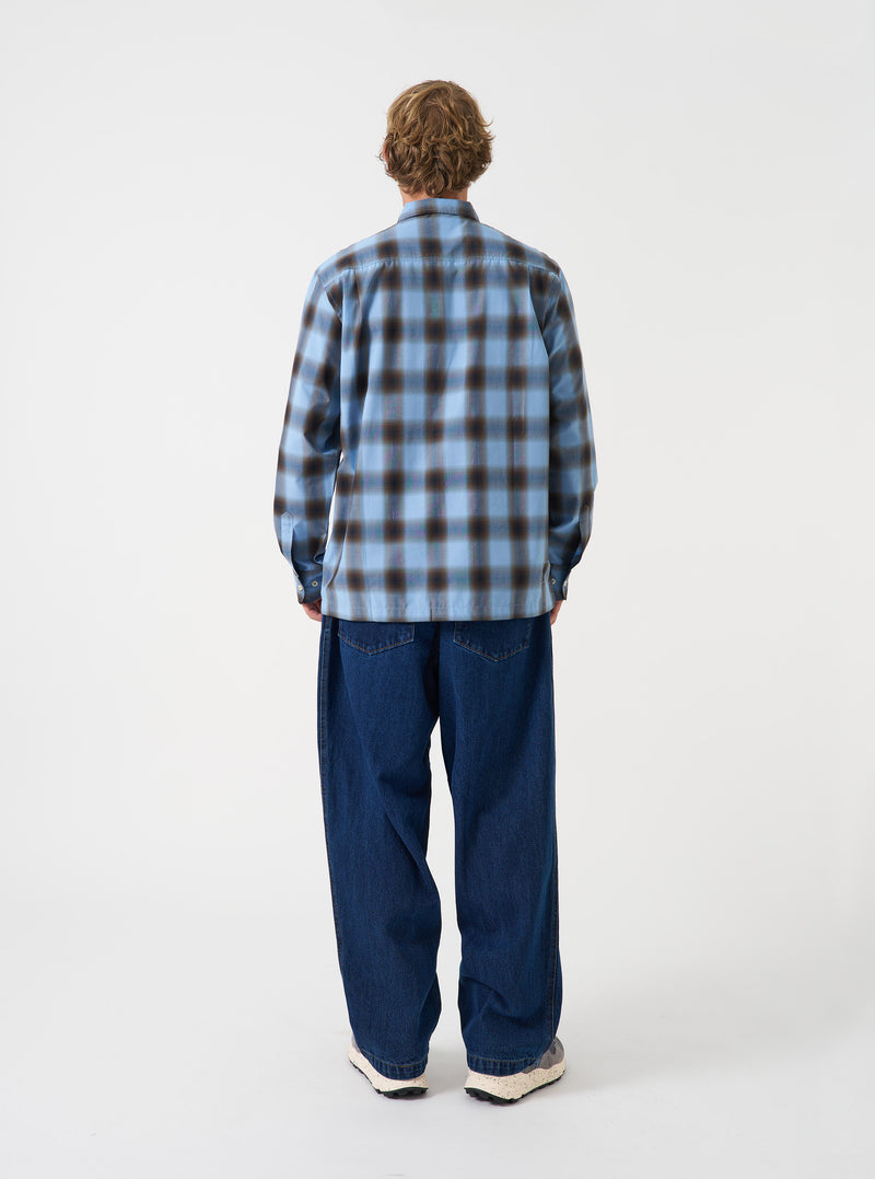 A back view of a model wearing the Universal Works Blue Check Charlie Check Attendant Shirt. The photo highlights the blue check patterned fabric across the back panel, relaxed shoulder construction, and straight hem set against a plain backdrop.
