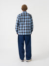 A back view of a model wearing the Universal Works Blue Check Charlie Check Attendant Shirt. The photo highlights the blue check patterned fabric across the back panel, relaxed shoulder construction, and straight hem set against a plain backdrop.