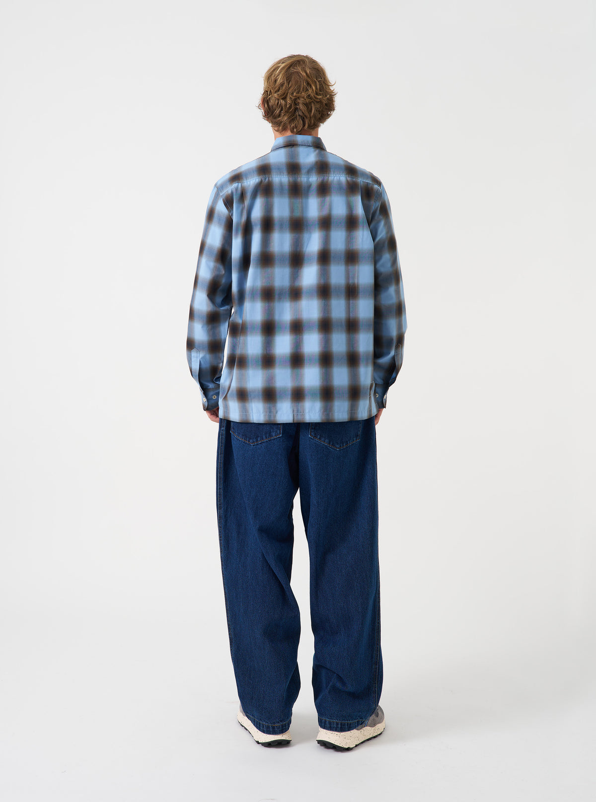 A back view of a model wearing the Universal Works Blue Check Charlie Check Attendant Shirt. The photo highlights the blue check patterned fabric across the back panel, relaxed shoulder construction, and straight hem set against a plain backdrop.