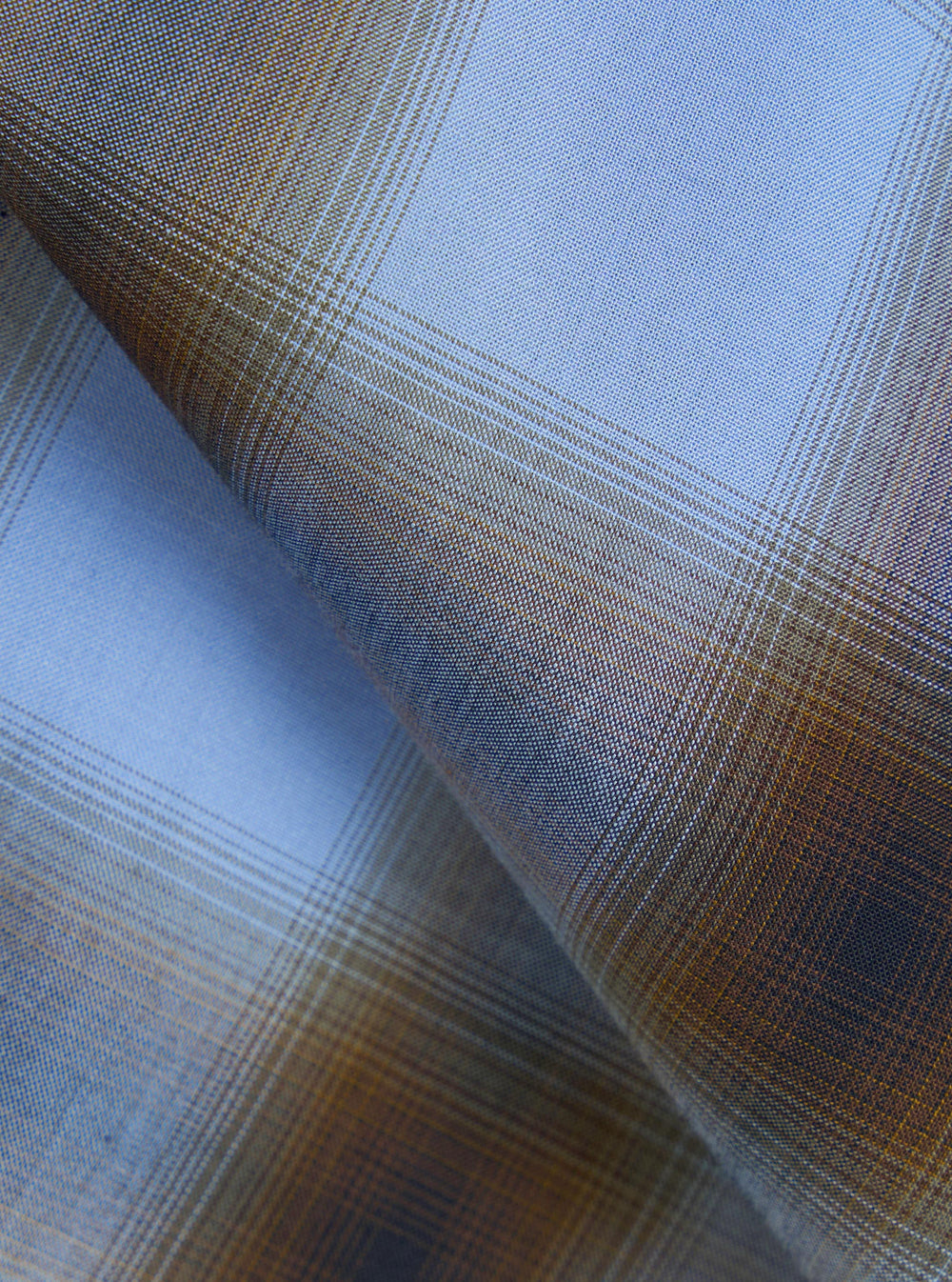 Close-up of the blue check fabric used in the Charlie Check Attendant Shirt. A fine woven cloth with a glen plaid pattern in pale blue, amber, and ochre. The folded fabric shows the weave texture and the contrast between the light blue ground and the warm brown overcheck lines.