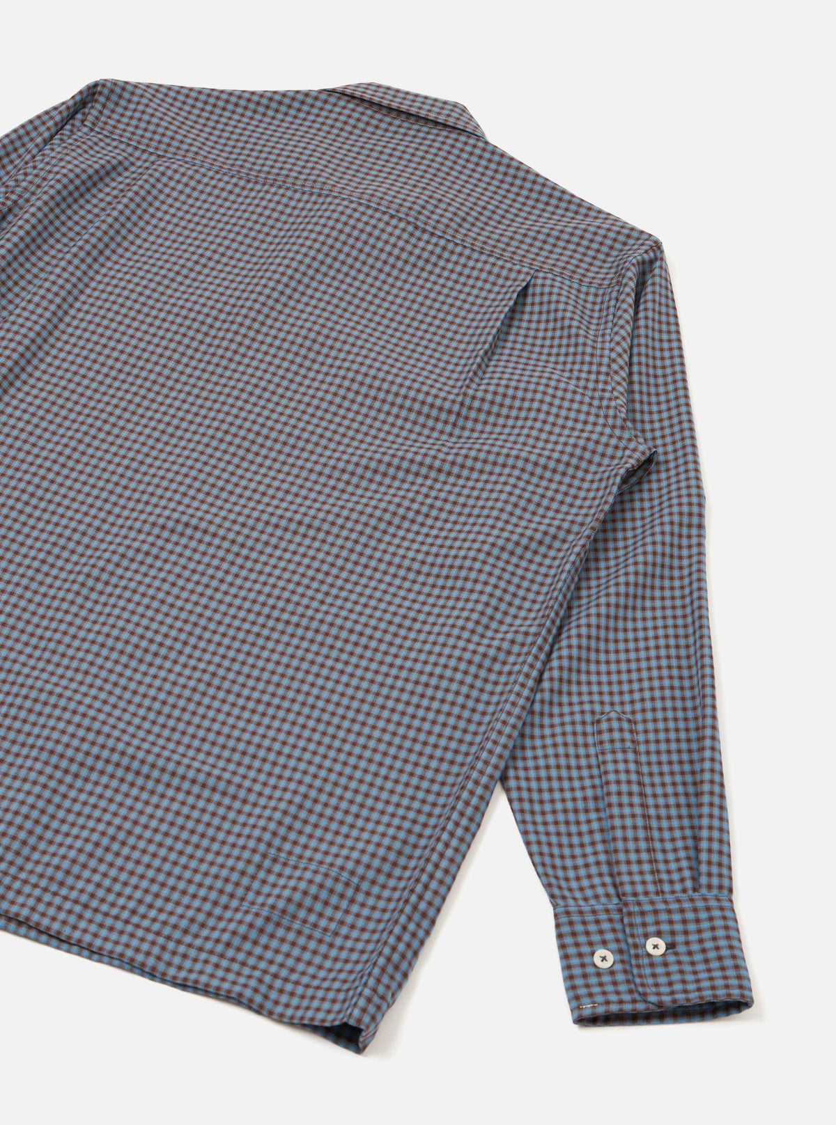 Flat lay of a blue and brown mini gingham check shirt on a white studio surface. The upper back, collar and one full sleeve are visible. The spread collar lies flat and the buttoned cuff features two small silver buttons. The woven check fabric has a fine texture throughout.