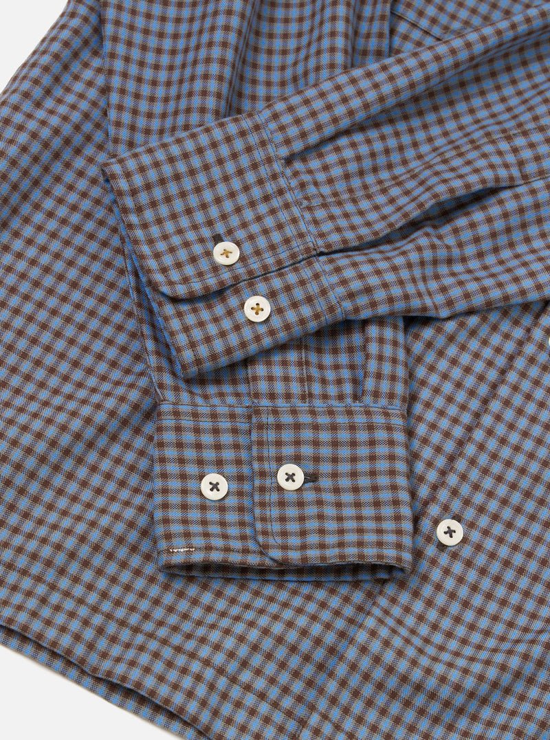 Flat lay detail shot of the blue and brown gingham check Attendant shirt on a white surface. The woven cotton fabric shows a fine two-colour check. Two buttoned barrel cuffs are visible, each fastened with two cream four-hole buttons. A chest patch pocket with a button tab is prominent in the foreground.