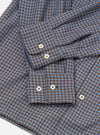 Flat lay detail shot of the blue and brown gingham check Attendant shirt on a white surface. The woven cotton fabric shows a fine two-colour check. Two buttoned barrel cuffs are visible, each fastened with two cream four-hole buttons. A chest patch pocket with a button tab is prominent in the foreground.