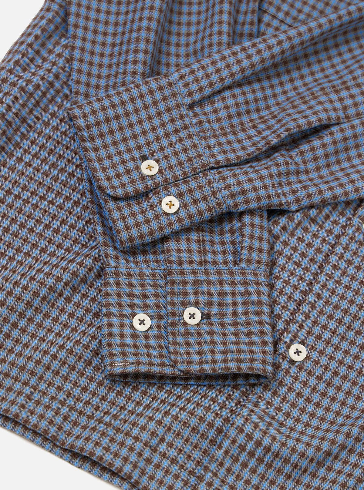 Flat lay detail shot of the blue and brown gingham check Attendant shirt on a white surface. The woven cotton fabric shows a fine two-colour check. Two buttoned barrel cuffs are visible, each fastened with two cream four-hole buttons. A chest patch pocket with a button tab is prominent in the foreground.