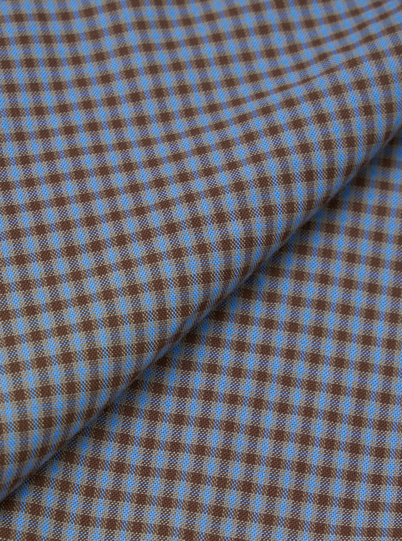 Close-up fabric detail of the blue and brown Parker check attendant shirt, showing a fine woven check pattern in mid blue and warm brown. The fabric is folded to reveal texture and weave. The tight micro-check creates a two-tone geometric effect across the woven surface.