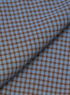 Close-up fabric detail of the blue and brown Parker check attendant shirt, showing a fine woven check pattern in mid blue and warm brown. The fabric is folded to reveal texture and weave. The tight micro-check creates a two-tone geometric effect across the woven surface.