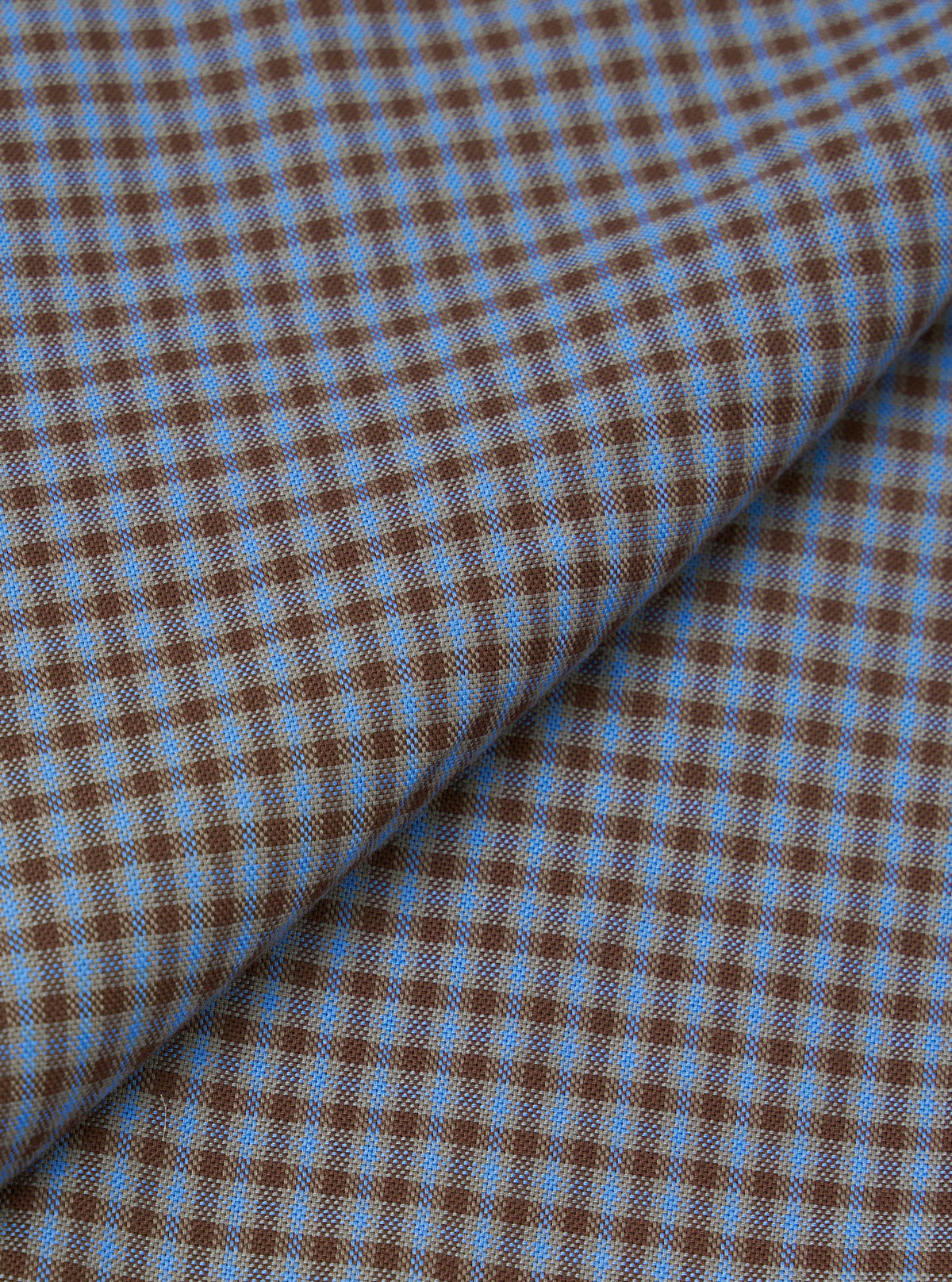 Close-up fabric detail of the blue and brown Parker check attendant shirt, showing a fine woven check pattern in mid blue and warm brown. The fabric is folded to reveal texture and weave. The tight micro-check creates a two-tone geometric effect across the woven surface.