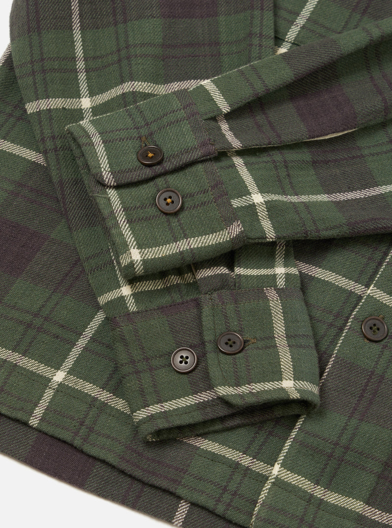 A close-up detail shot of the Universal Works Olive Washed Check L/S Utility Shirt against a plain background, highlighting the olive green check woven cotton texture, buttoned chest pocket, and fine stitching construction.