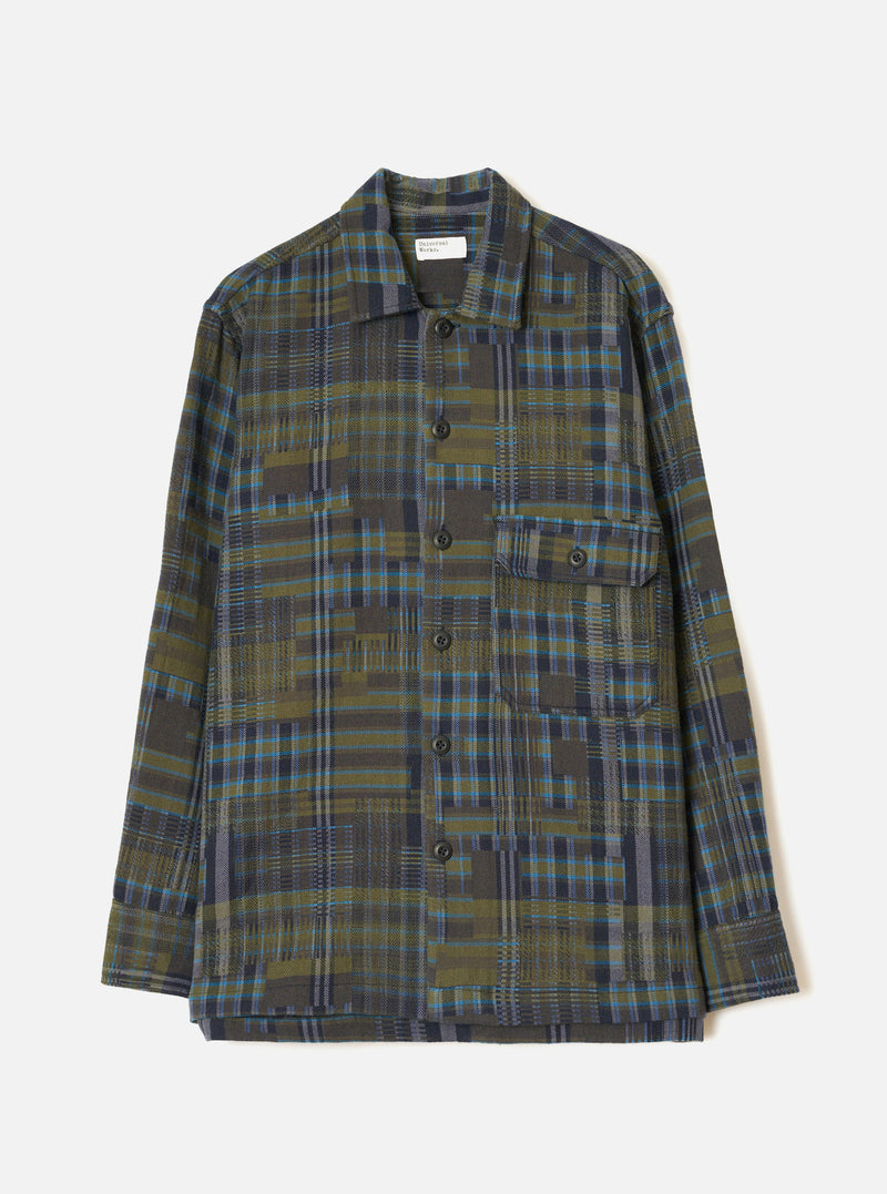 Olive shadows check fatigue shirt laid flat on a light grey seamless backdrop. The shirt features a camp collar, a single patch chest pocket with a buttoned flap, and a five-button placket with dark buttons. Long sleeves with plain cuffs. The fabric is a patchwork-style plaid in olive, navy, and teal with a textured weave.