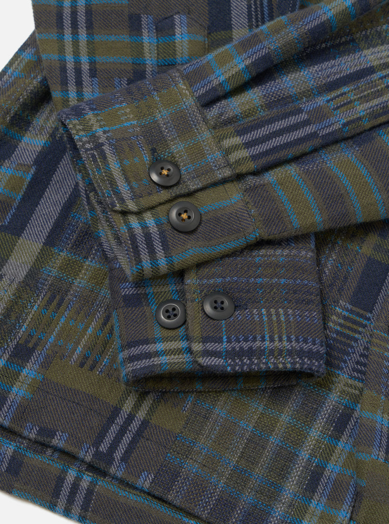 Close-up flat lay of an olive shadows check fatigue shirt in brushed flannel. The shot focuses on the sleeve and cuff, showing four dark horn-style buttons: two on the placket and two on the buttoned cuff. The plaid weave combines olive, navy, grey and teal on a white studio backdrop.