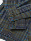 Close-up flat lay of an olive shadows check fatigue shirt in brushed flannel. The shot focuses on the sleeve and cuff, showing four dark horn-style buttons: two on the placket and two on the buttoned cuff. The plaid weave combines olive, navy, grey and teal on a white studio backdrop.