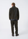 A back view of a model wearing the Olive Flock Chambray Warmus Jacket set against a plain backdrop. The shot showcases the smooth olive green cotton fabric across the back, clean shoulders, and relaxed fit.