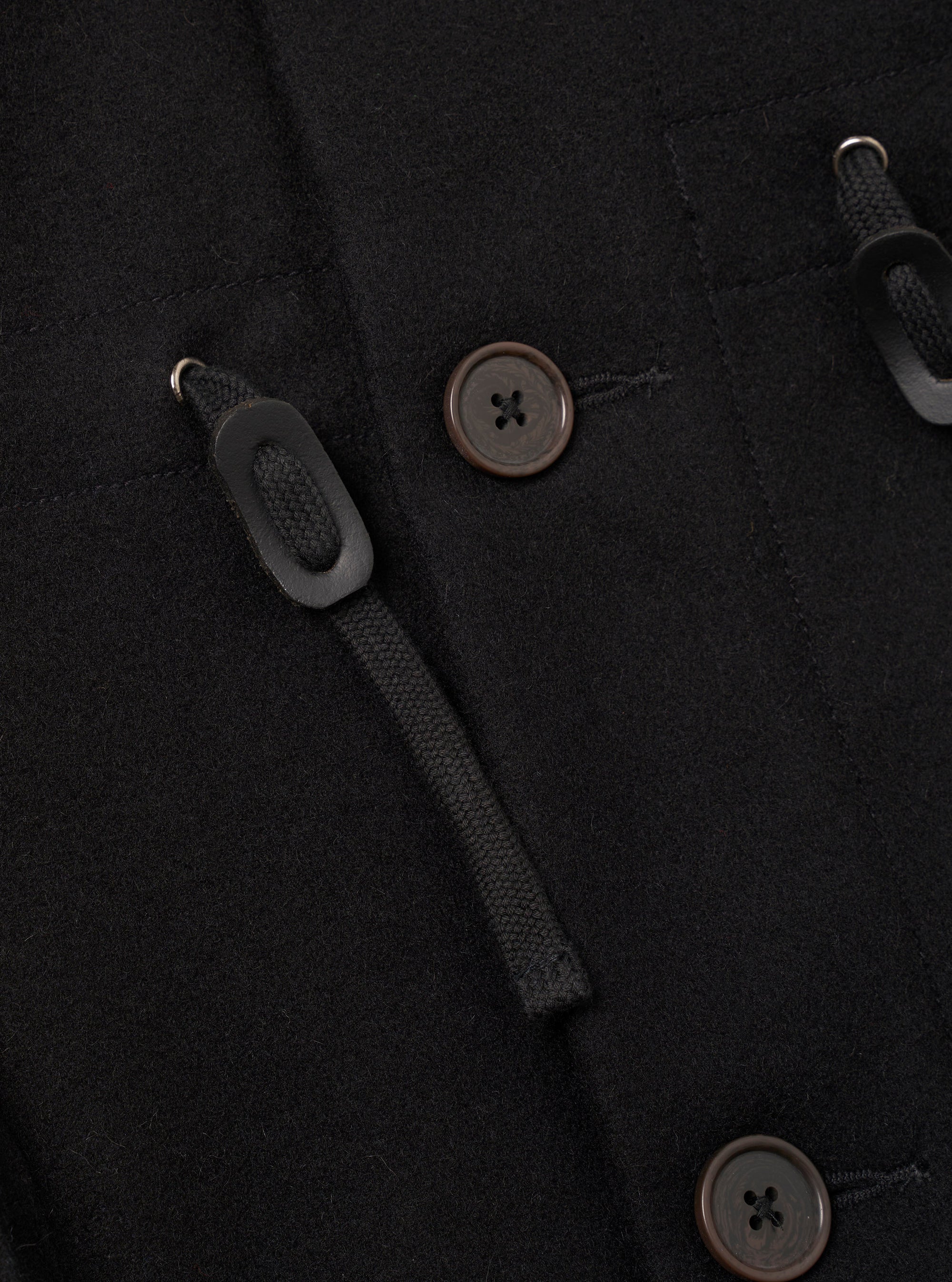 Close-up of the black Mowbray Boston Parka in a brushed wool-blend fabric. Two dark brown four-hole buttons are visible alongside toggle-and-loop duffle fastenings: braided black cord threaded through a metal ring and secured with a matte black oblong toggle. A patch pocket seam is visible at the top of the frame.