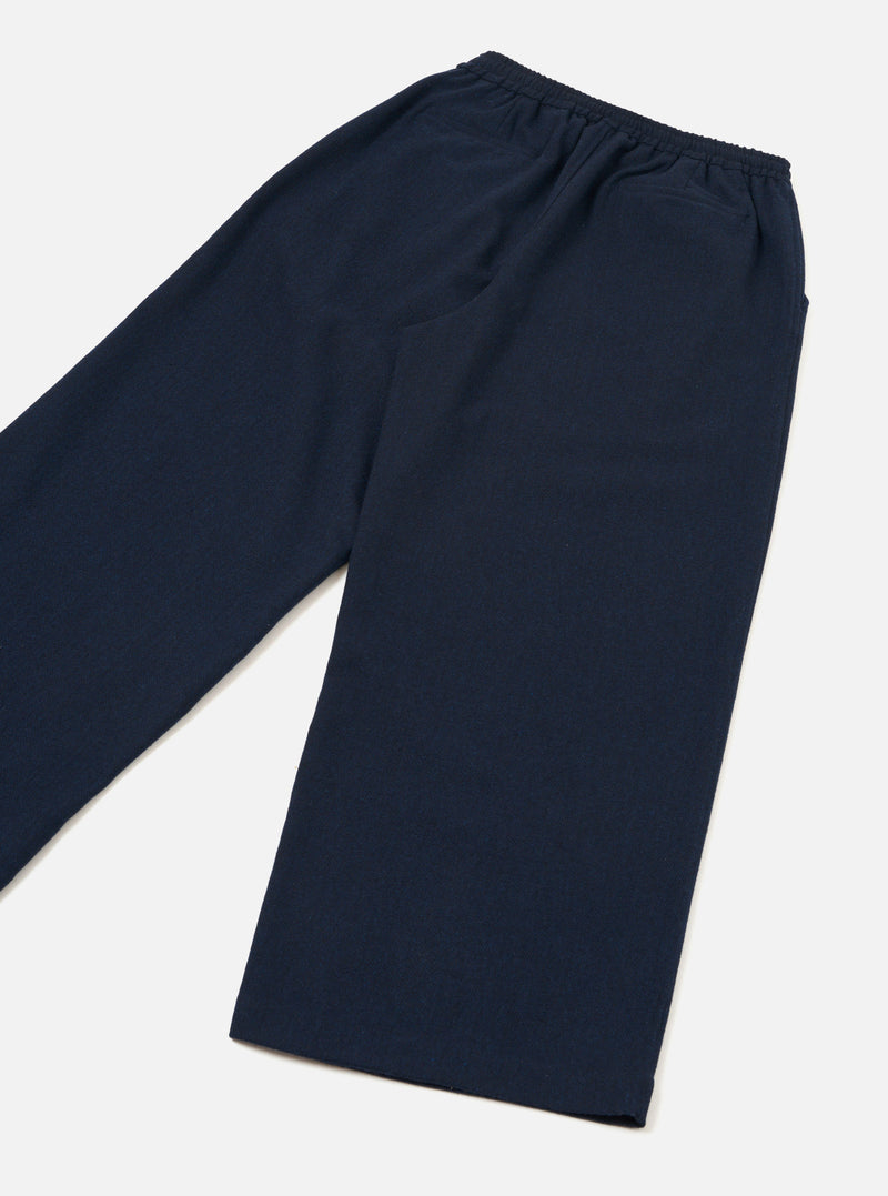 The Navy Wool Herringbone Gillespie Pant is laid flat on a light grey surface, captured at an angle. It displays a gathered elasticated waistband, side pocket opening, subtle wool herringbone fabric texture, and wide legs tapering to plain hems.