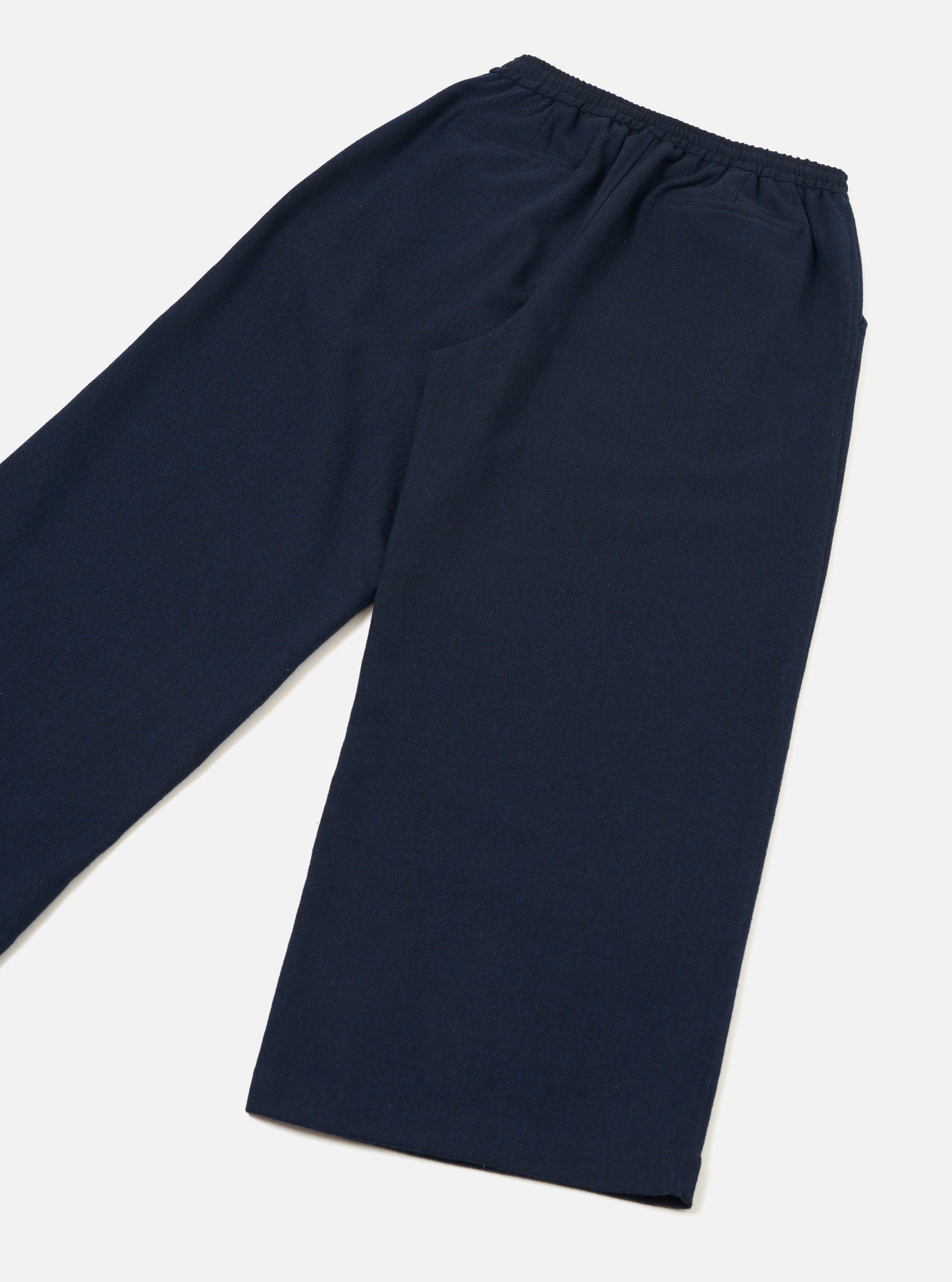 Navy wool herringbone trousers laid flat on a light grey surface, shot at an angle. A fully elasticated waistband sits at the top with visible gather. The wide leg tapers slightly to a plain unfinished hem. A side pocket opening is visible on the right. The plain-weave fabric shows a subtle texture across the surface.