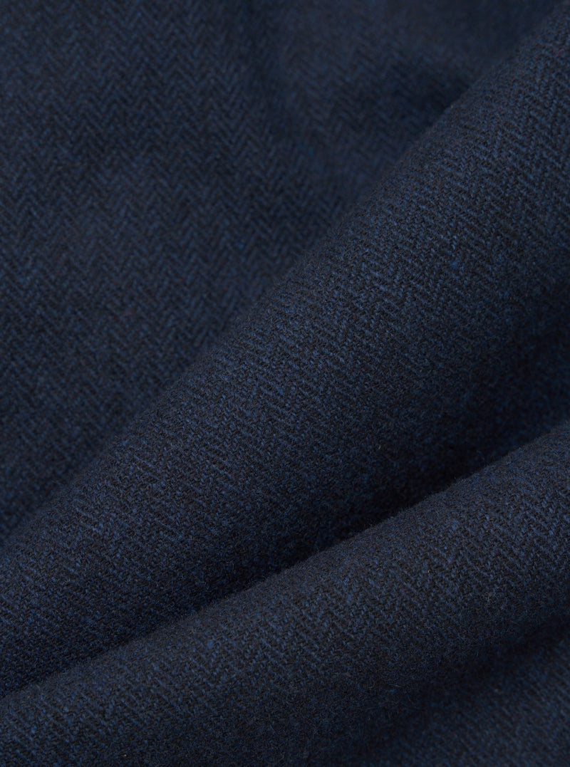 Close-up of navy wool herringbone fabric, showing the diagonal zigzag weave pattern in deep navy and dark charcoal tones with subtle blue flecks. The cloth is folded to highlight its texture and weight. No garment construction details are visible.