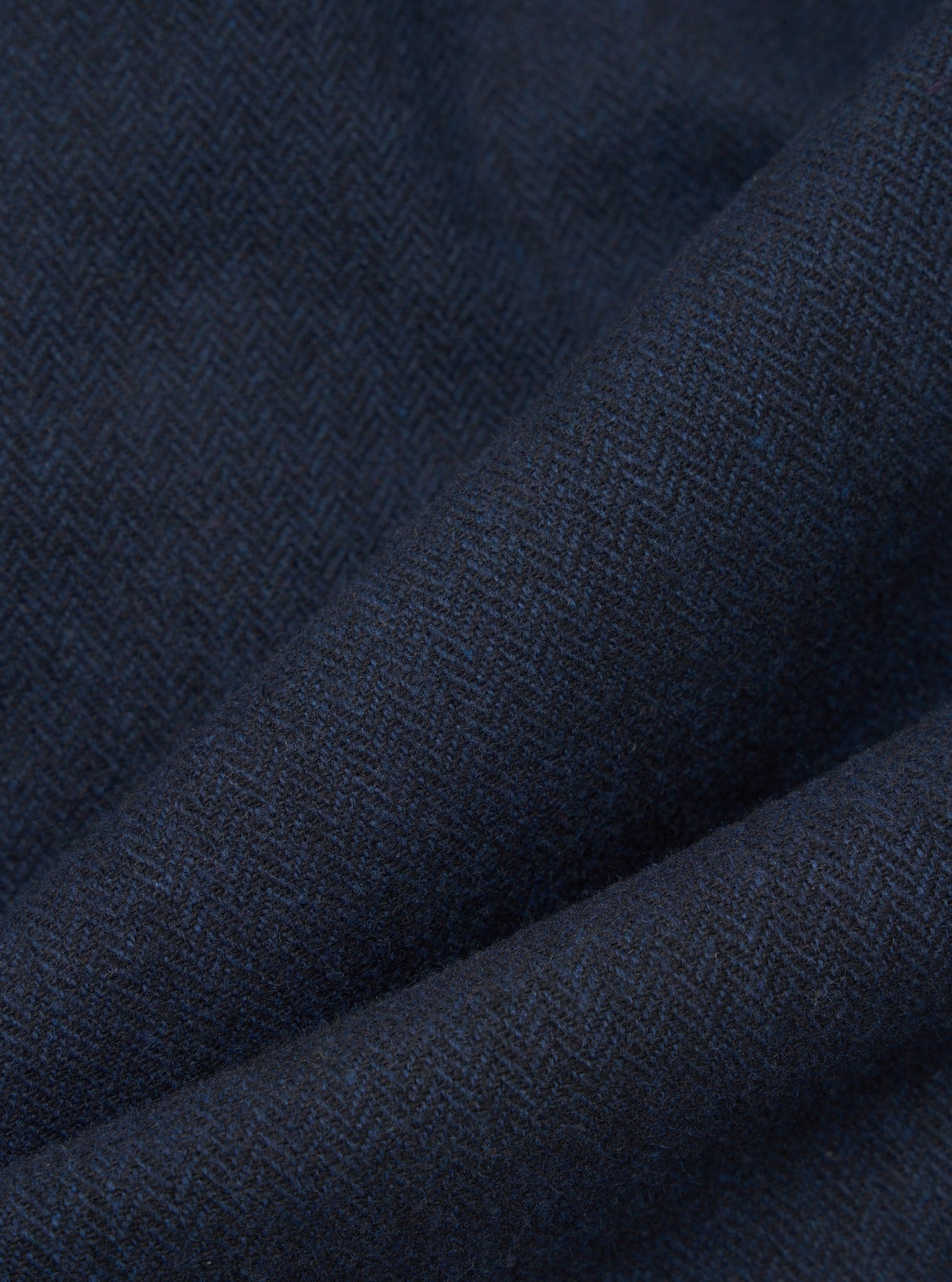 Close-up of navy wool herringbone fabric, showing the diagonal zigzag weave pattern in deep navy and dark charcoal tones with subtle blue flecks. The cloth is folded to highlight its texture and weight. No garment construction details are visible.