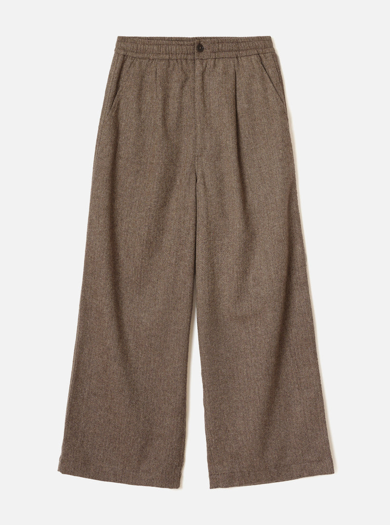Brown wool herringbone wide-leg trousers laid flat on a plain white background. The elasticated waistband features a single dark button at centre front. Slanted side pockets sit below the waistband. The fabric shows a classic herringbone weave in brown and cream tones. Full-length legs with a generous wide leg and plain hem.