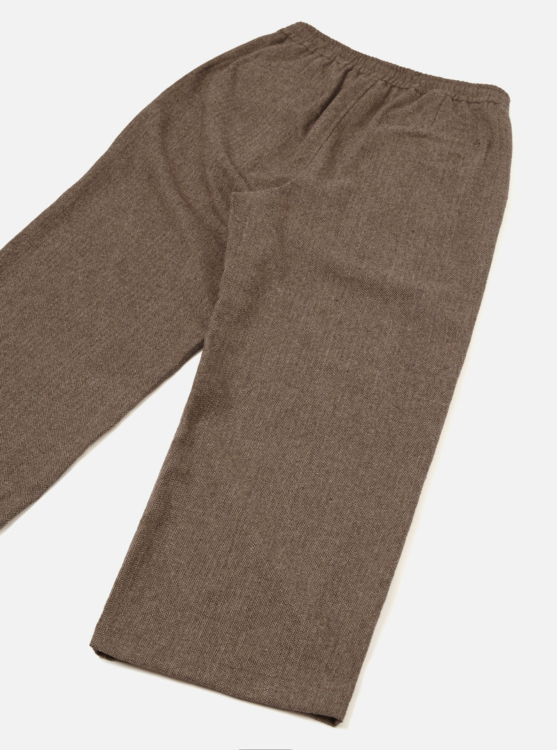 Brown wool herringbone Gillespie trousers laid flat on a white studio surface, angled diagonally. The herringbone weave is clearly visible across the fabric. An elasticated waistband sits at the top with a patch pocket on the rear right. The straight-leg cut tapers slightly to an unfinished hem.