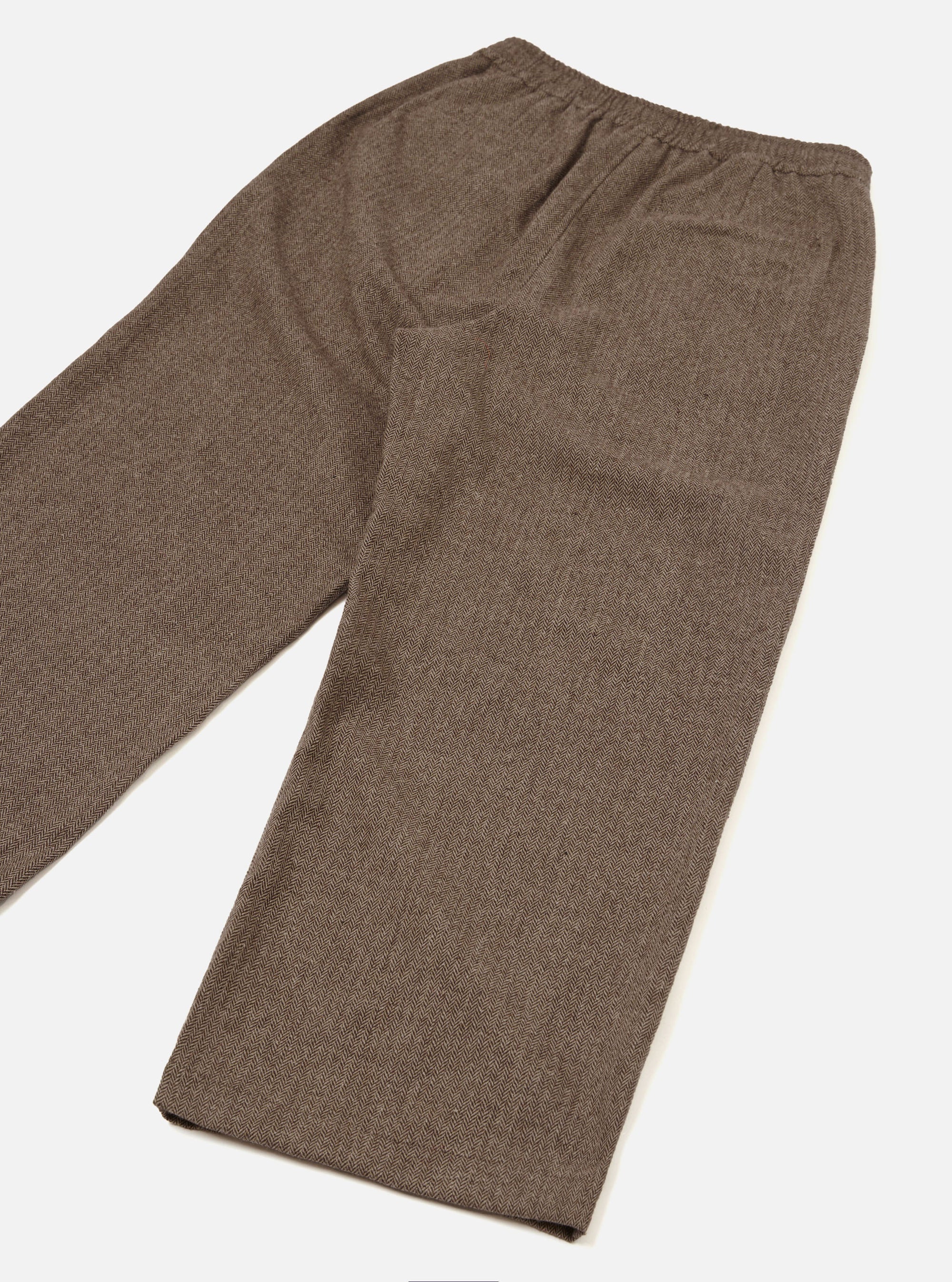 Brown wool herringbone Gillespie trousers laid flat on a white studio surface, angled diagonally. The herringbone weave is clearly visible across the fabric. An elasticated waistband sits at the top with a patch pocket on the rear right. The straight-leg cut tapers slightly to an unfinished hem.