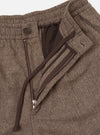 Close-up detail shot of the brown wool herringbone Gillespie pant on a white studio backdrop. The fly is open, showing a metal zip, a dark brown button at the waistband, a zip-fastened welt pocket on the fly flap, and a dark brown lining. The herringbone weave is clearly visible across the fabric.
